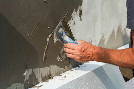 Close-up of detail of worker hand with trowel applying glue on plastered white house wall doing...