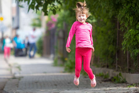 Cute small funny smiling toothless girl in pink casual clothing with long blond pony tail jumping and having fun on blurred bright sunny street outdoors background. Happy careless childhood concept.の写真素材