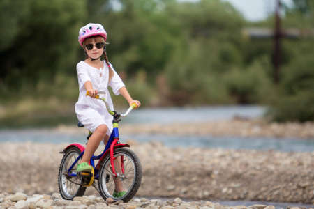 Cute young girl in white clothing, sunglasses with long braids wearing pink safety helmet riding child bicycle on pebbled river bank on blurred green summer background. Outdoors activity concept.の写真素材