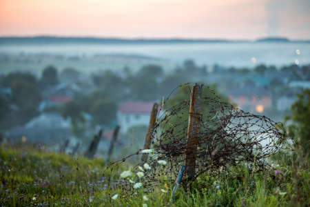 Old big barbed wire coiled on rusty pole, broken garden fence on grassy blooming hill on misty light...