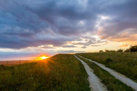 Beautiful landscape at sunset or sunrise, narrow ground road stretching through green grassy...