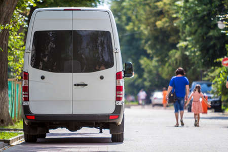 Back view of white passenger medium size commercial luxury minibus van parked n shadow of green tree on summer city street  with blurred silhouettes of pedestrians and cars under green trees.の写真素材