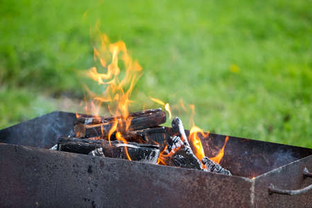 Brightly burning in metal box logs firewood for barbecue outdoor on sunny day. Orange high flame and white smoke on blurred blue sky and green grass background. Camping, safety and tourism concept.の写真素材