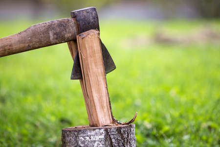 Close-up of old sharp dirty weathered iron ax cutting through chunk of firewood on tree trunk outdoors on blurred green grassy sunny blooming summer meadow bokeh background.の写真素材