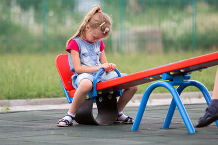 Playground conflicts. Small cute young blond child girl sits moody, angry and offended on see- saw swing on bright green blurred background. Joys and sorrows of childhood, children tantrums concept.の写真素材