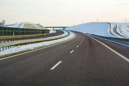 Modern wide smooth empty asphalt highway stretching to horizon under high bridge turning right past distant forest at day. Speed, comfortable journey and professional road building concept.の写真素材