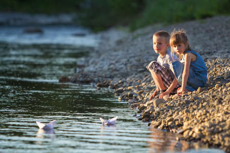 Two smiling blond children, boy and girl playing with white paper boats on river bank on bright summer blurred blue background. Joys and games of happy childhood and outdoors activities concept.の写真素材