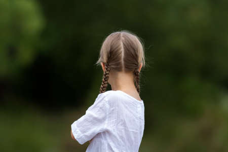 Back view portrait of cute fashionable young girl head with long blond braids in white dress on blurred sunny summer green outdoors copy space bokeh background.の写真素材