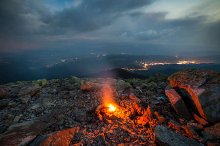 Beautiful summer night in mountains. Bright white yellow fire burning on rocky mountain top under...