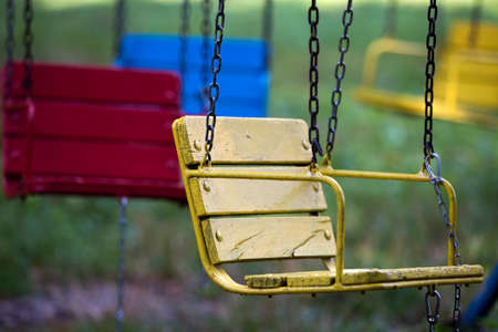 Close-up of empty wooden painted blue, yellow, vintage seats chairs of multi-colored carousel hanging on chains outdoor in city amusement park on sunny day on bright green blurred background.の写真素材