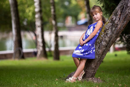 Adorable pretty young child girl with beautiful long blond hair in fashionable blue dress leaning on growing tree trunk on park green grassy lawn, lake water surface and blurred trees background.の写真素材