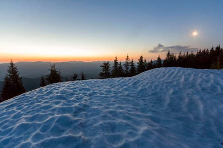 Fantastic view of evening in winter mountains. Dark green tops of pine trees behind snowy hill and first bright star in quiet blue sky with orange glow over horizon. Breathtaking beauty of nature.の写真素材