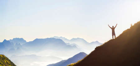 Man on peak of mountain. Emotional scene. Young man with backpack standing with raised hands on top...
