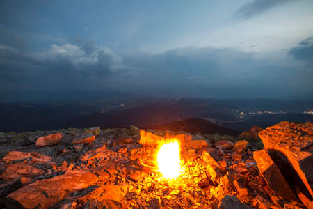 Beautiful summer night in mountains. Bright white yellow fire burning on rocky mountain top under blue cloudy sky, dark mountain ridge stretching to horizon and small road lights below in valley.の写真素材