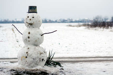 Big funny traditional primitive smiling snowman with bucket hat on white snowy field winter landscape, blurred black trees and blue sky copy space background. Merry Christmas and happy New Year.の写真素材