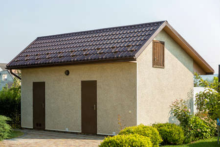 New detached small barn, workshop or utility building with brown shingle roof, two doors and attic on clean paved yard on summer sunny day. Well maintained fenced propert, real estate concept.のeditorial素材
