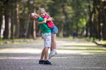 Two cute young funny smiling children, girl and boy, brother holding sister in his arms, having fun on blurred bright sunny park alley green trees bokeh background. Loving siblings relations concept.の写真素材