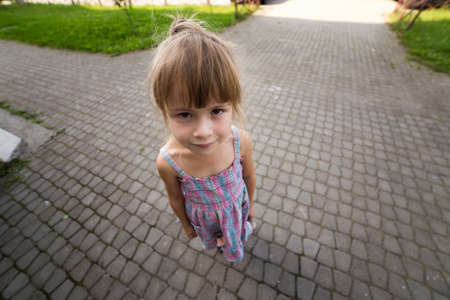 Close-up portrait, angled view from above of pretty young little blond pale unhappy moody friendless child girl looking sadly in camera on blurred sunny outdoors background. Children problems concept.の写真素材