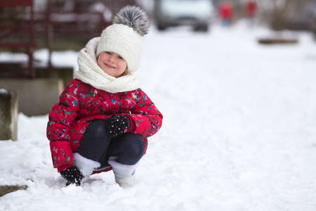 Portrait of cute little young funny smiling child girl in nice warm clothing playing in snow having fun on winter cold day on white bright blurred copy space background. Outdoors activity and games.の写真素材