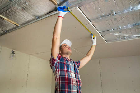 Young handsome man in casual clothing takes measurement of drywall suspended ceiling connected to...