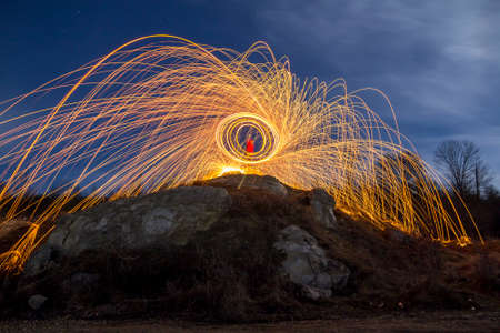 Long exposure shot of man standing on rocky hill spinning steel wool in circle making firework showers of bright yellow glowing sparkles on blue night sky background. Light painting art concept.のeditorial素材