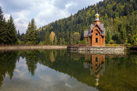Summer view of beautiful wooden orthodox chapel in ecological area on small artificial island on background of green spruce mountain and bright blue sky reflected in clear lake water on sunny day.のeditorial素材