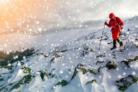 Tourist hiker in bright red clothing with walking sticks descending dangerous rocky mountain slope covered with snow on stormy cloudy sky copy space background.の写真素材