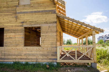 Part of unfinished wooden ecological traditional cottage of natural lumber materials with steep roof frame and attached terrace with decorative railing under construction in green neighborhood.の写真素材