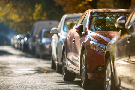 Long row of different shiny cars parked along empty roadside on sunny autumn day on blurred green...