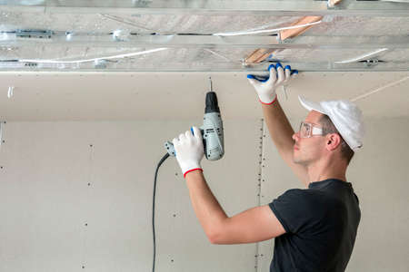 Young man in goggles fixing drywall suspended ceiling to metal frame using electrical screwdriver on...
