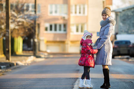Back view of young slim attractive woman mother and small child girl daughter in warm clothing walking holding hands on sunny winter day on blurred urban background.の写真素材