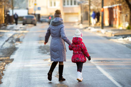 Back view of young slim attractive woman mother and small child girl daughter in warm clothing walking together holding hands crossing slippery street on sunny winter day on blurred urban background.の写真素材