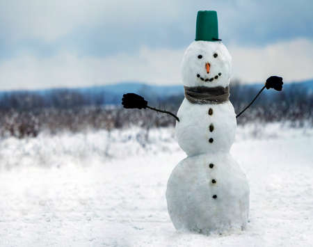 Big smiling snowman with bucket hat, scarf and gloves on white snowy field winter landscape, blurred black trees and blue sky copy space background. . Merry Christmas and happy new year greeting card.の写真素材
