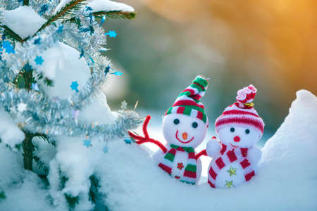Two small funny toys baby snowman in knitted hats and scarves in deep snow outdoors near pine tree branch. Happy New Year and Merry Christmas greeting card.の写真素材