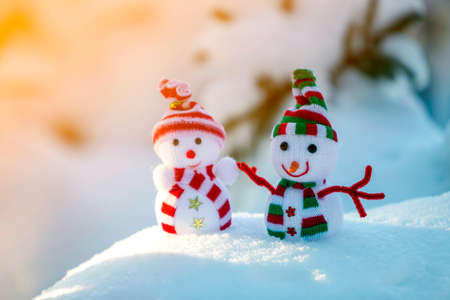 Two small funny toys baby snowman in knitted hats and scarves in deep snow outdoors near pine tree branch. Happy New Year and Merry Christmas greeting card.の写真素材