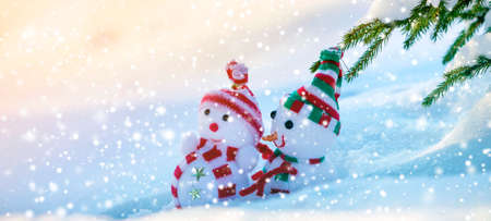 Two small funny toys baby snowman in knitted hats and scarves in deep snow outdoors near pine tree branch. Happy New Year and Merry Christmas greeting card.の写真素材