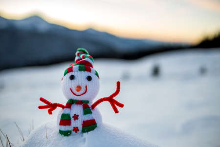 Small funny toy baby snowman in knitted hat and scarf in deep snow outdoor on blurred snow covered mountains landscape background. Happy New Year and Merry Christmas greeting card theme.の写真素材
