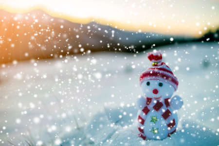 Small funny toy baby snowman in knitted hat and scarf in deep snow outdoor on blurred mountains landscape and falling big snowflakes background. Happy New Year and Merry Christmas greeting card theme.の写真素材