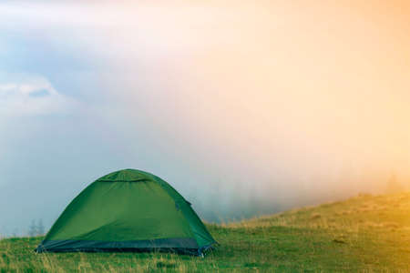 Small tourist tent on grassy mountain hill on fogy tree tops and clear blue sky before sunrise copy space background. Summer camping in mountains at dawn. Tourism, hiking and beauty of nature concept.の写真素材