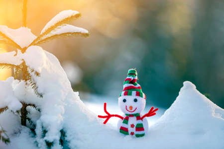 Small funny toy baby snowman in knitted hat and scarf in deep snow outdoors on bright blue and white...