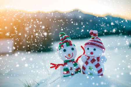 Two small funny toys baby snowman in knitted hats and scarves in deep snow outdoors on blurred mountains landscape background. Happy New Year and Merry Christmas greeting card theme.の写真素材