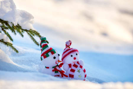 Two small funny toys baby snowman in knitted hats and scarves in deep snow outdoors near pine tree branch. Happy New Year and Merry Christmas greeting card.の写真素材