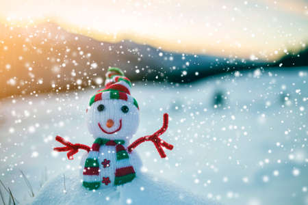 Small funny toy baby snowman in knitted hat and scarf in deep snow outdoor on blurred mountains landscape and falling big snowflakes background. Happy New Year and Merry Christmas greeting card theme.の写真素材