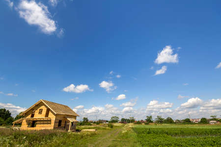 New wooden ecological not finished cottage from natural materials under construction in green field on distant village and blue sky copy space background. Old traditions and modern building concept.の写真素材