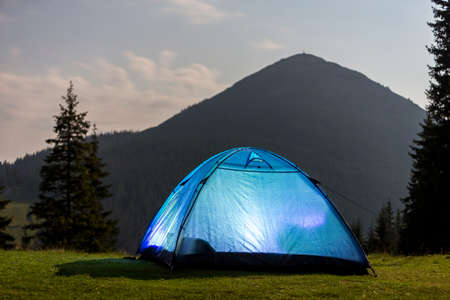 Tourist hikers bright blue tent on green grassy forest clearing among tall pine trees under clear morning sky on distant foggy woody mountain background. Tourism, summer camping in mountains at dawn.の写真素材