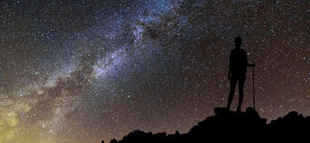 Back view of young slim backpacker tourist girl on rocky mountain top on dark night starry sky and foggy mountain range panorama background. Tourism, traveling and climbing concept.の写真素材