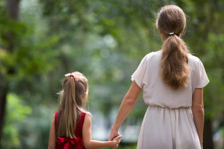 Back view of small blond long-haired child girl with slender slim mother in fashionable dresses walking together holding hands on warm sunny day outdoors on blurred colorful sunny bokeh background.の写真素材