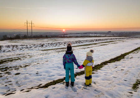 Back view of two young children in warm clothing standing in frozen snow field holding hands on copy space background of setting sun and clear blue sky.の写真素材