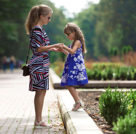 Profile full-length portrait of young blond long-haired attractive woman and small child girl in sunglasses and fashionable dresses holding hands on sunny park alley. Happy family relations concept.の写真素材
