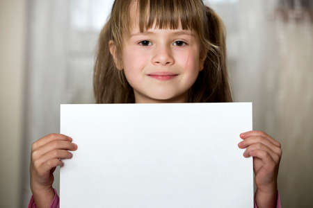 Happy smiling cute child girl holding white copy space sheet of drawing paper. Art education, creativity, advertisement concept.の写真素材